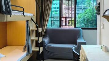 Quarto com cama de solteiro e bicama, sofá, janela com cortinas e mesa com luminária.