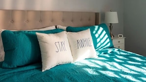 A bed with teal bedding and two pillows with the words "STAY. AWAY." The bed has a tufted headboard and is positioned next to a nightstand with a lamp. - Desert Lily - 6 Pools, Pickle Ball & Tennis Courts, 2 Patios (Palm Springs)