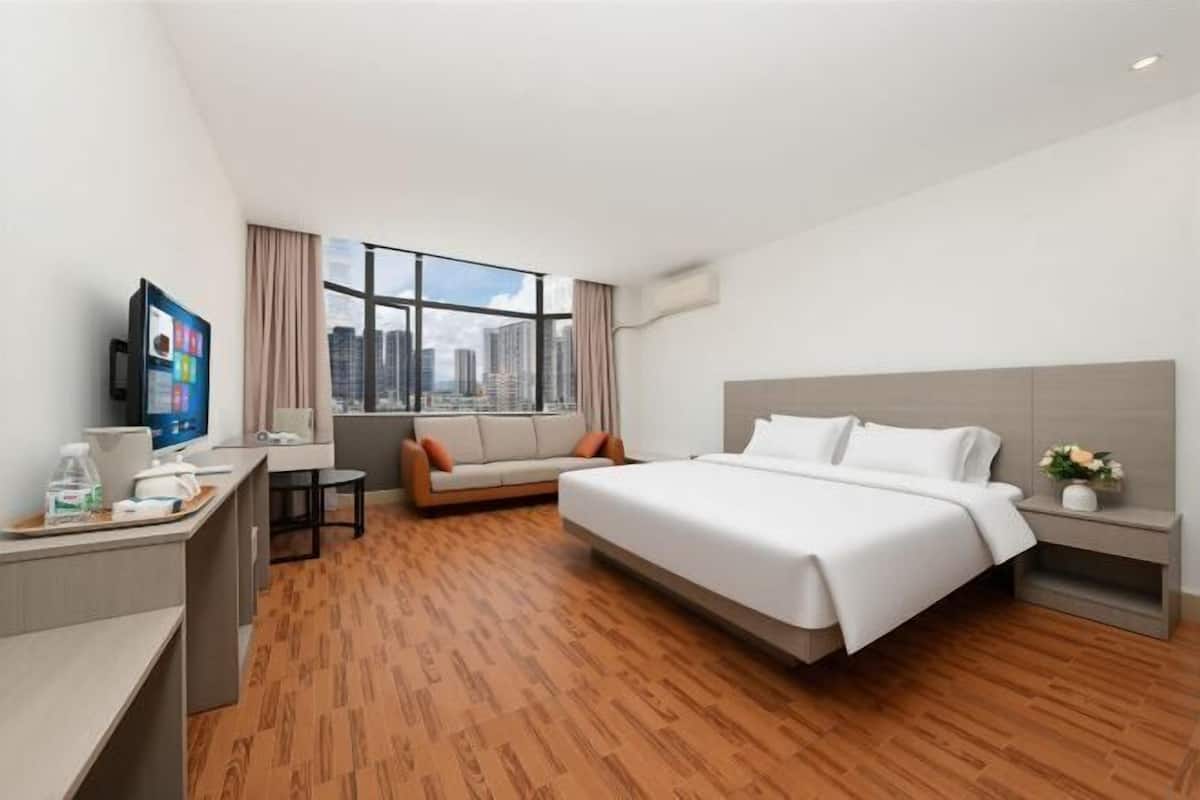 A hotel room with a bed, a desk with a computer, a sofa, a window with a city view, and a small table with a vase of flowers.