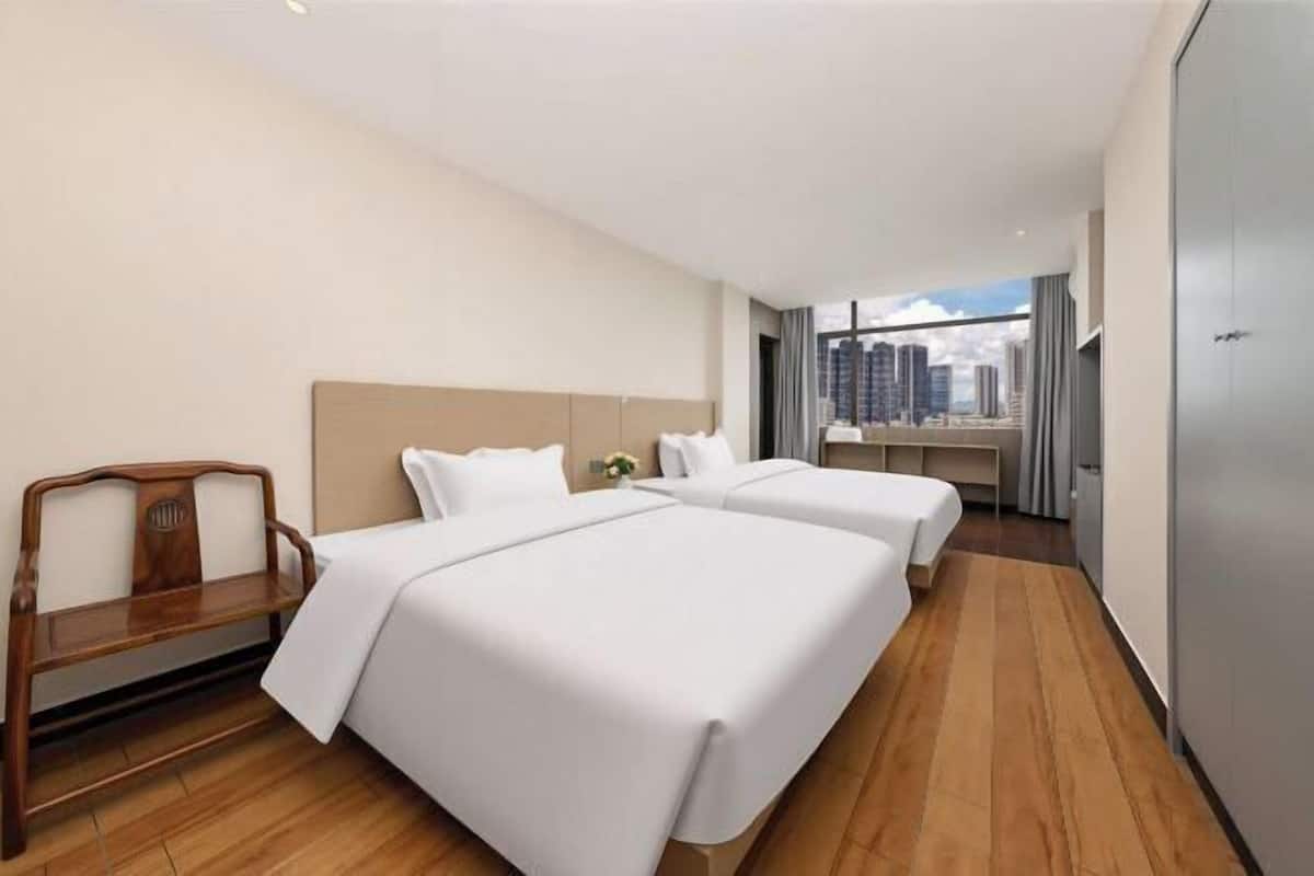 A hotel room with two beds, a wooden bench, a large window with a city view, and a wardrobe.