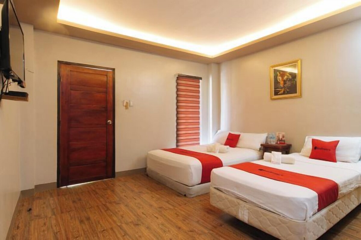 Quarto de hotel com duas camas, porta de madeira, janela com persianas, quadro emoldurado na parede e mesa de cabeceira com abajur.