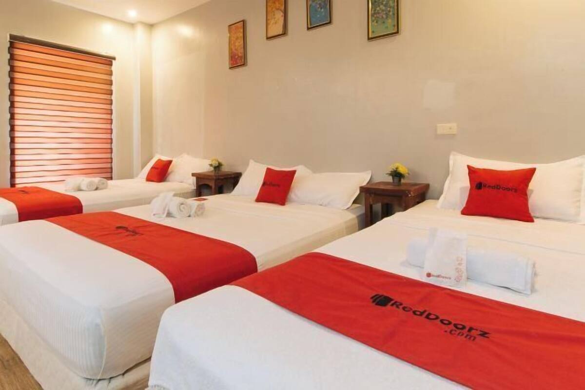 Ein Hotelzimmer mit drei Betten, jeweils mit roter Bettdecke und passenden Kissen, sowie einem Fenster mit Jalousien.