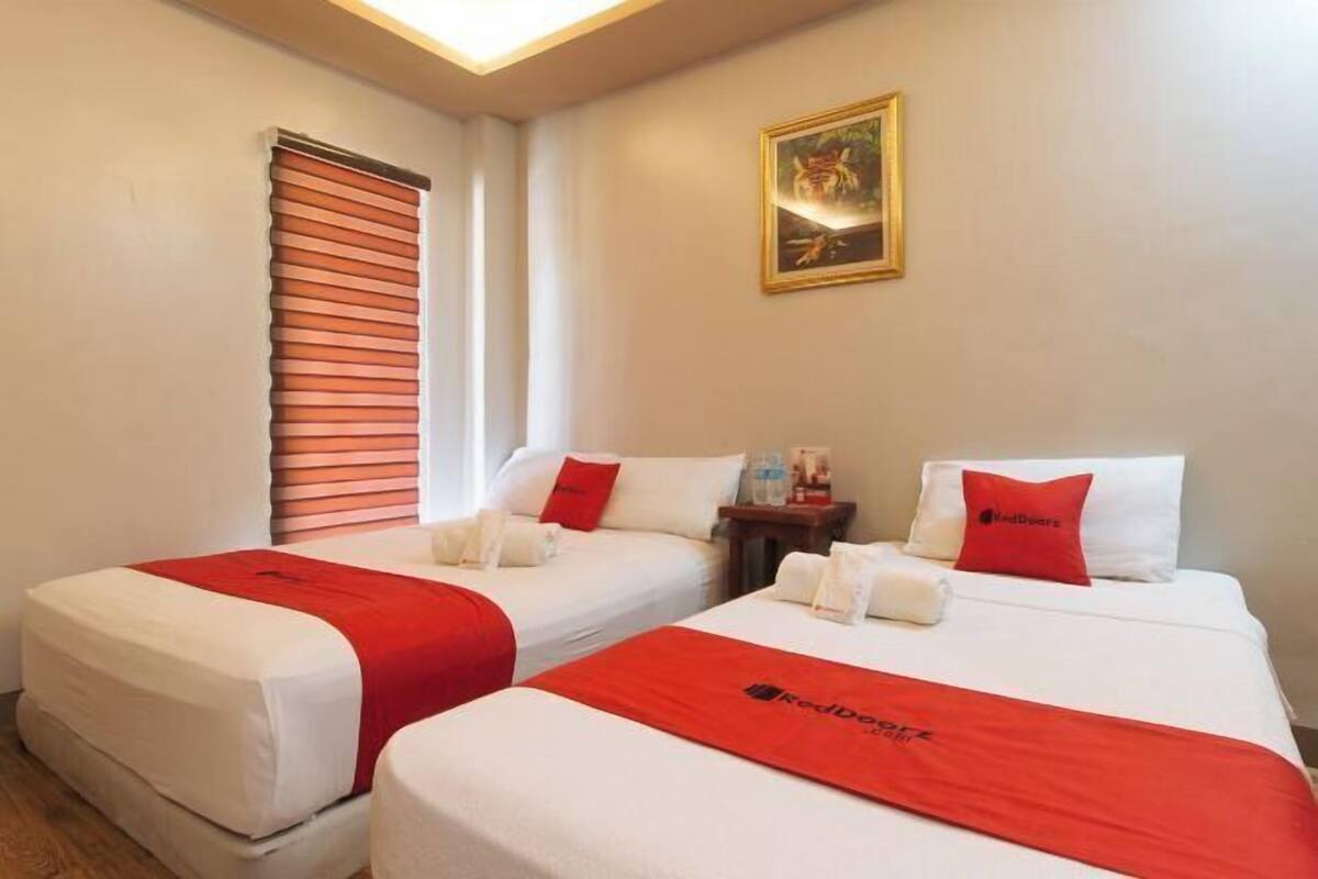 Ein Hotelzimmer mit zwei Betten, jeweils mit roter Decke und Kissen, einem Bild an der Wand und einem Fenster mit Jalousien.