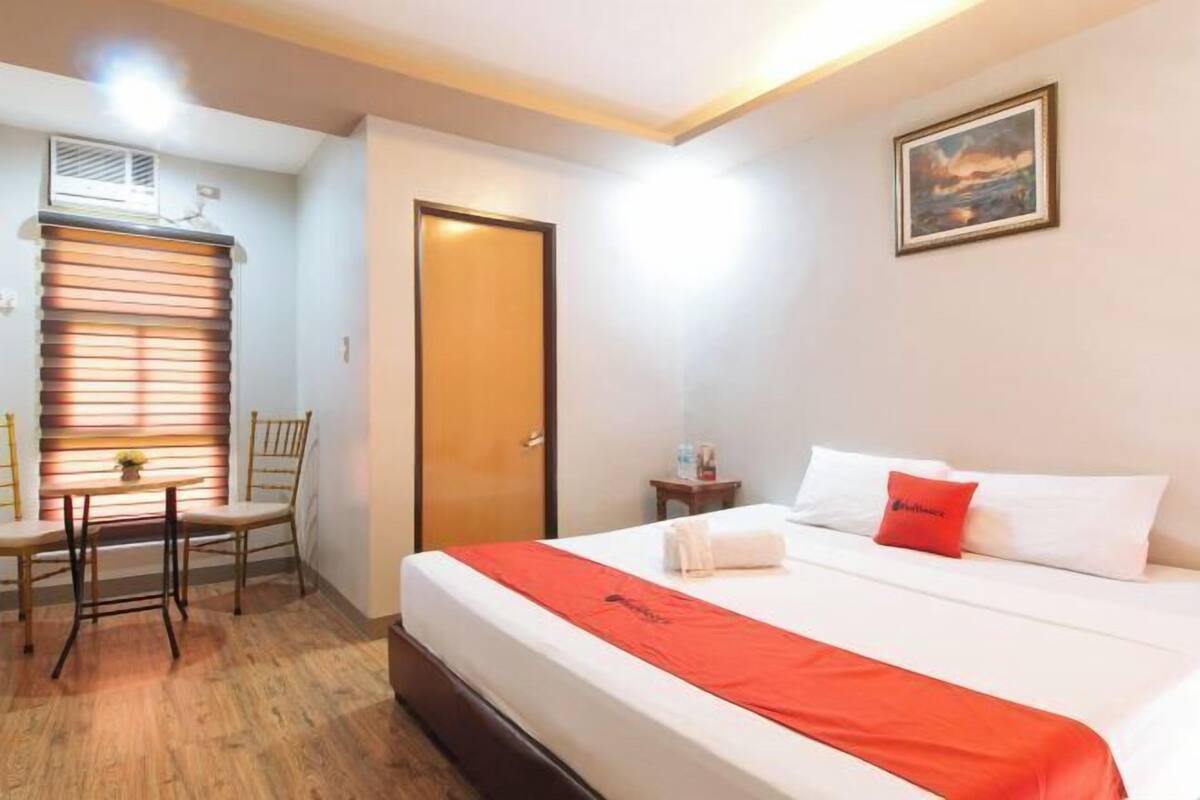 Ein Hotelzimmer mit einem Bett, einem Schreibtisch mit Stühlen, einem Fenster mit Jalousien, einer Tür und einem Bild an der Wand.