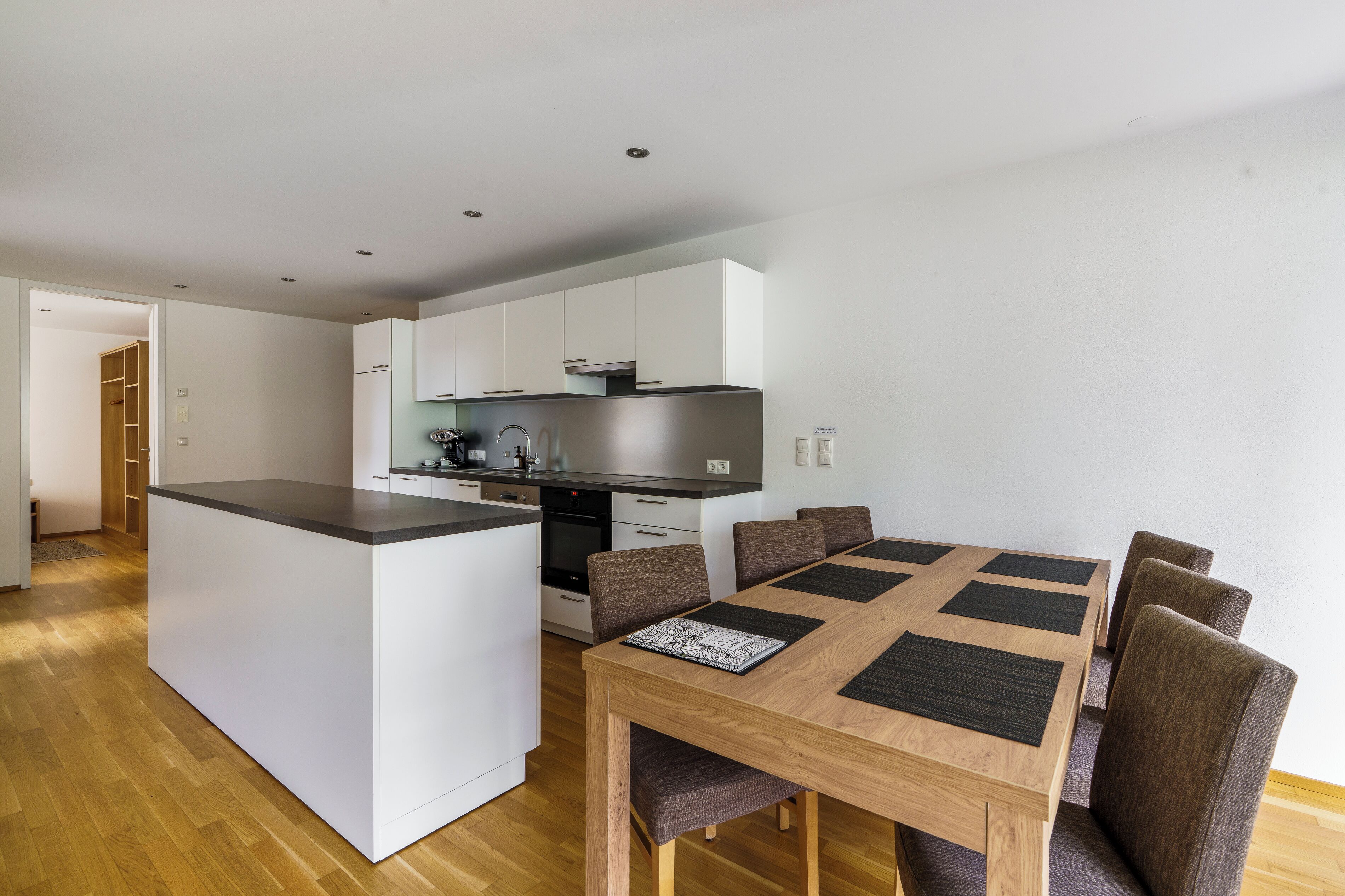 Una cocina moderna con armarios blancos, una isla central y un comedor con mesa y sillas de madera.
