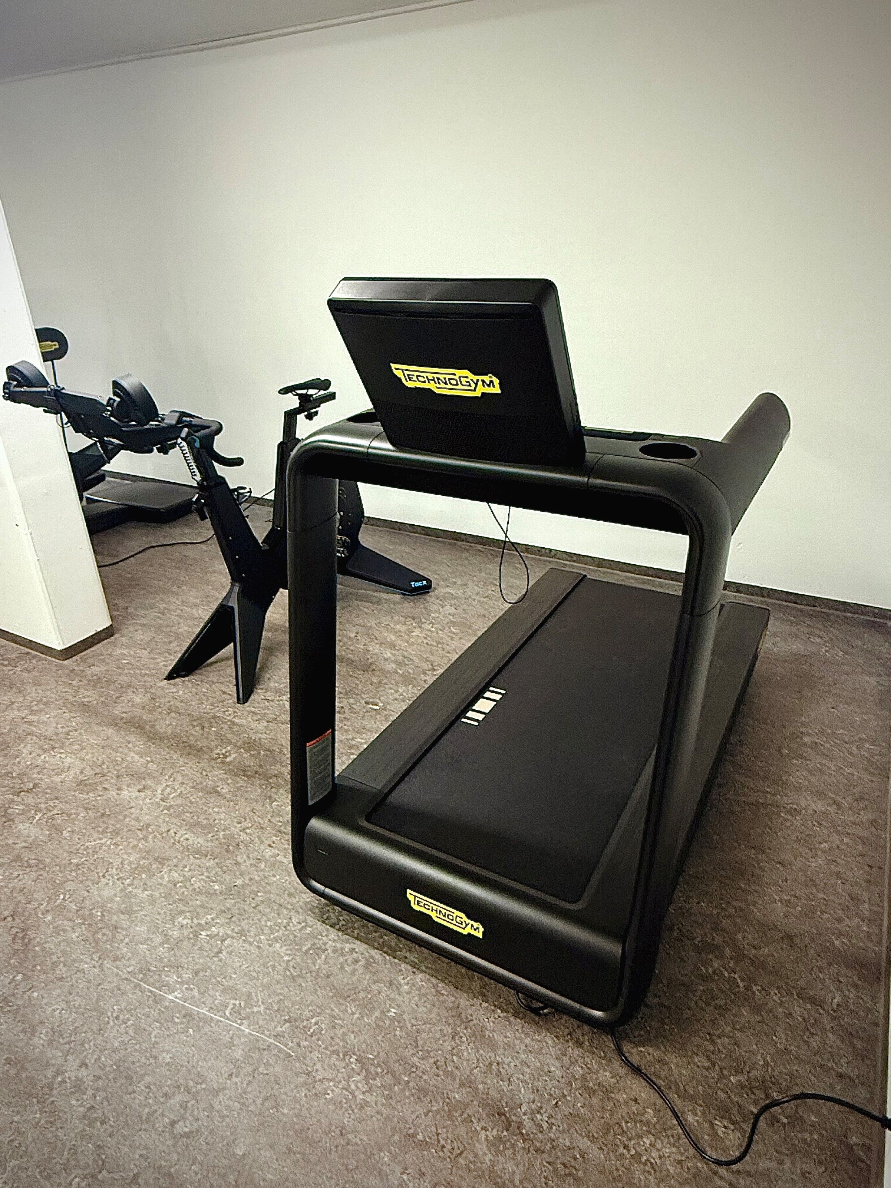 A treadmill with a laptop on top, a stationary bike, and a white wall in the background.