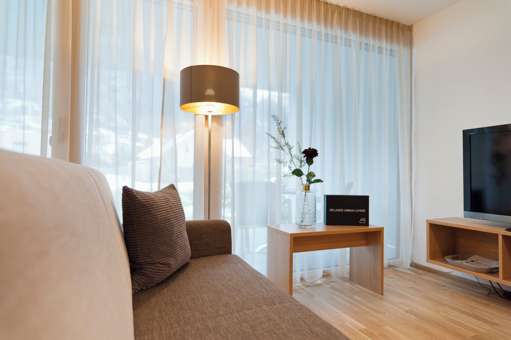 Una sala de estar moderna con un sofá, una lámpara, un jarrón con flores, un televisor y un letrero que dice 'RELAXED URBAN LIVING'.