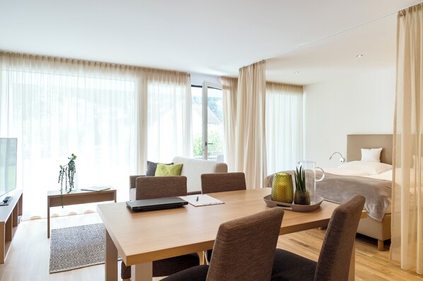 A modern hotel room with a wooden dining table, a bed with a white headboard, and a large window with sheer curtains. - Relaxed Urban Living (Dornbirn)