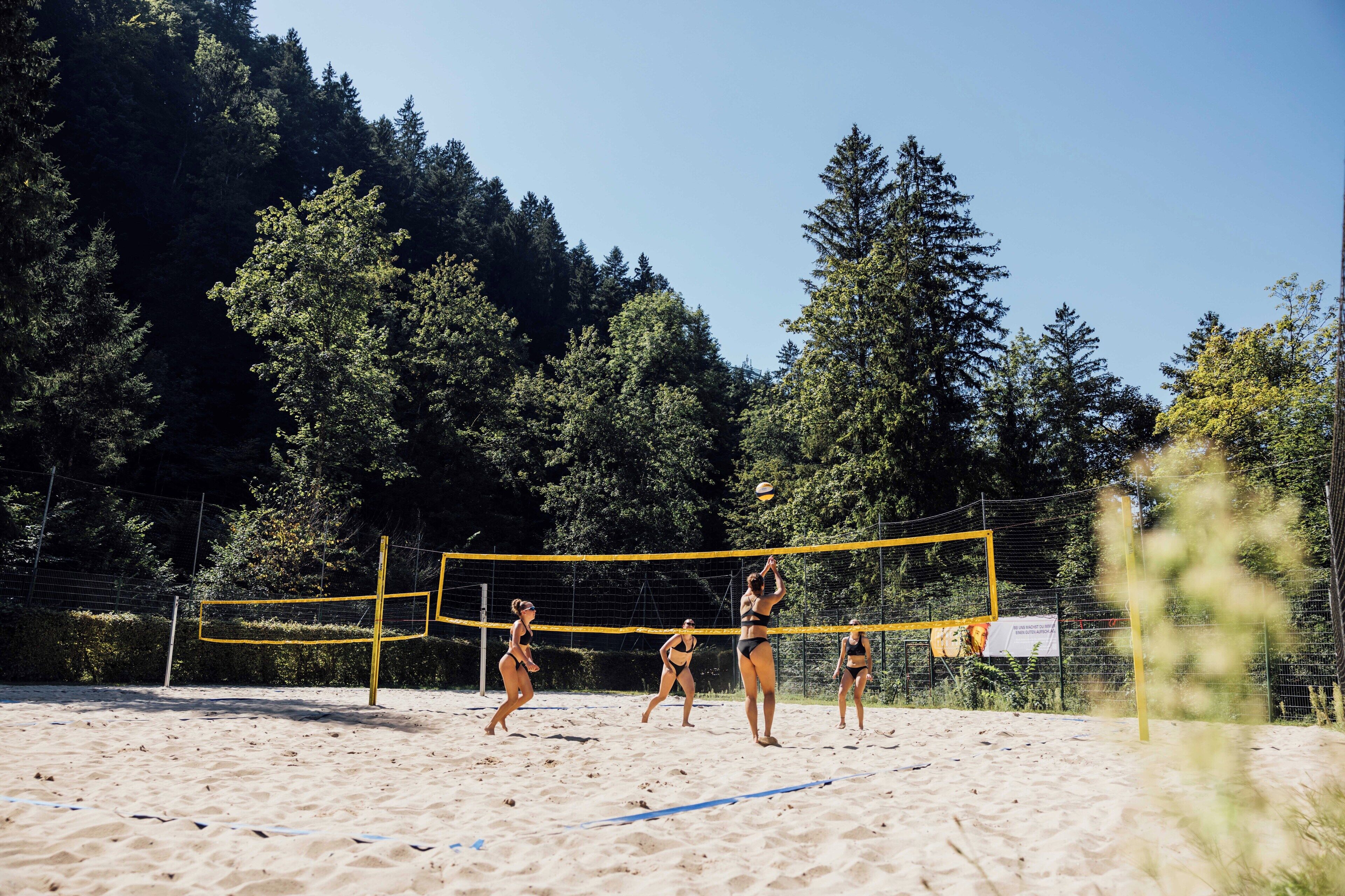 A beach volleyball court with a net, surrounded by sand and trees.