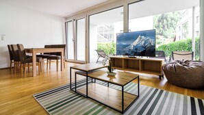A modern living room with a wooden coffee table, a TV on a wooden stand, and a large window offering a view of greenery and buildings. - Relaxed Urban Living (Dornbirn)