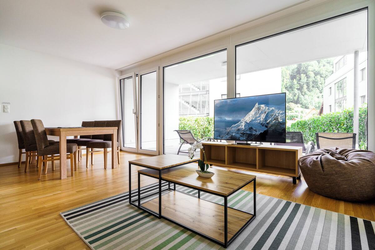 A modern living room with a wooden coffee table, a TV on a wooden stand, and a large window offering a view of greenery and buildings.