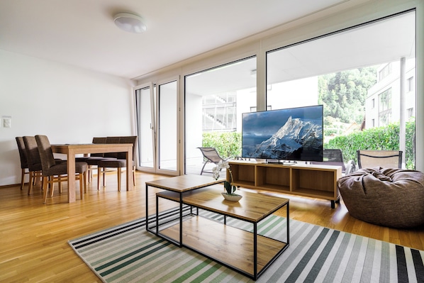 A modern living room with a wooden coffee table, a TV on a wooden stand, and a large window offering a view of greenery and buildings. - Relaxed Urban Living (Dornbirn)