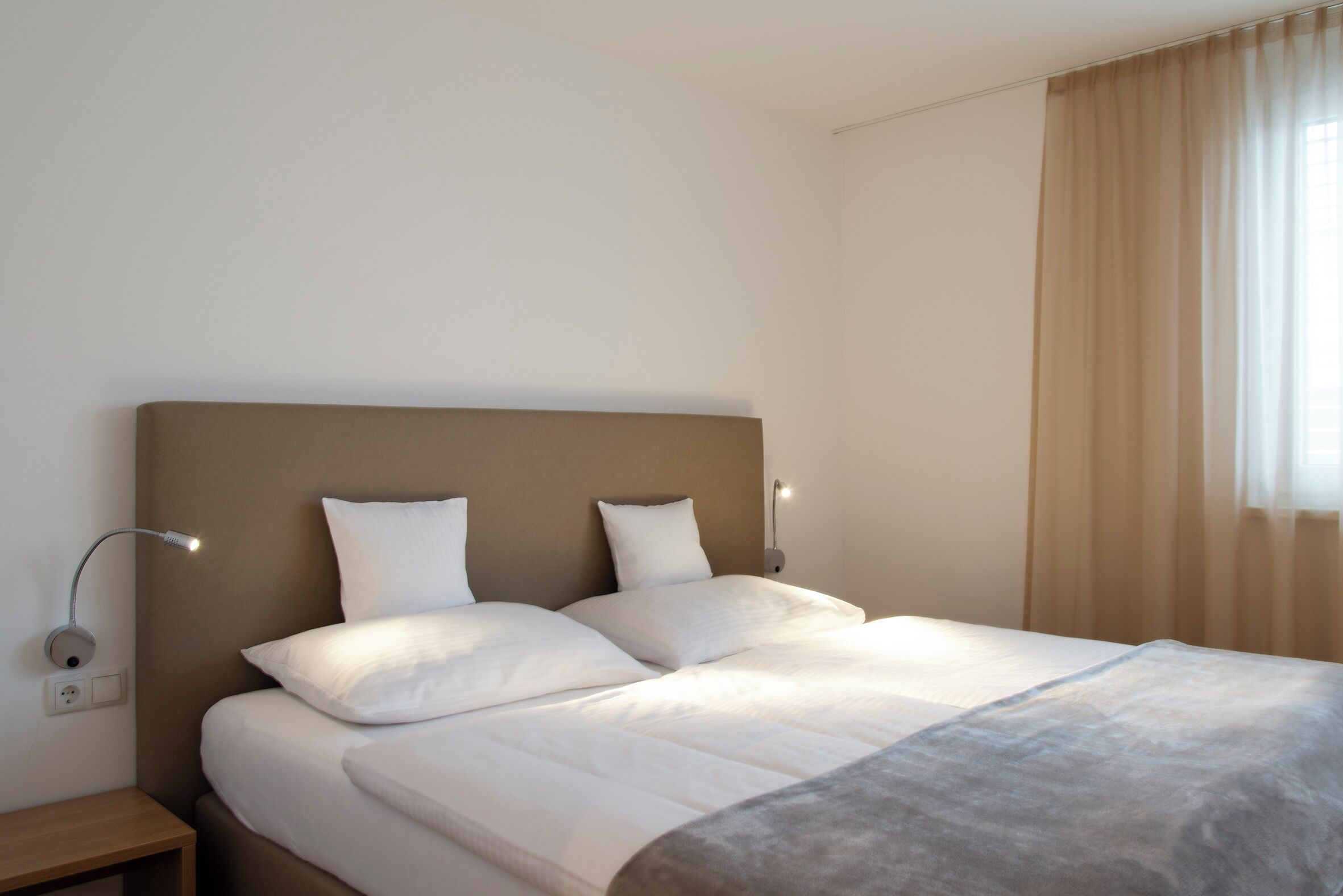 Una cama bien tendida con cabecera, dos lámparas de noche y una ventana con cortinas.