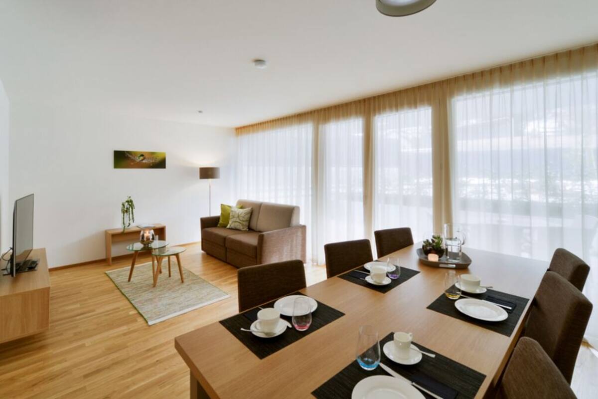 A modern living room with a wooden dining table set for four, a sofa, a TV, and a plant.