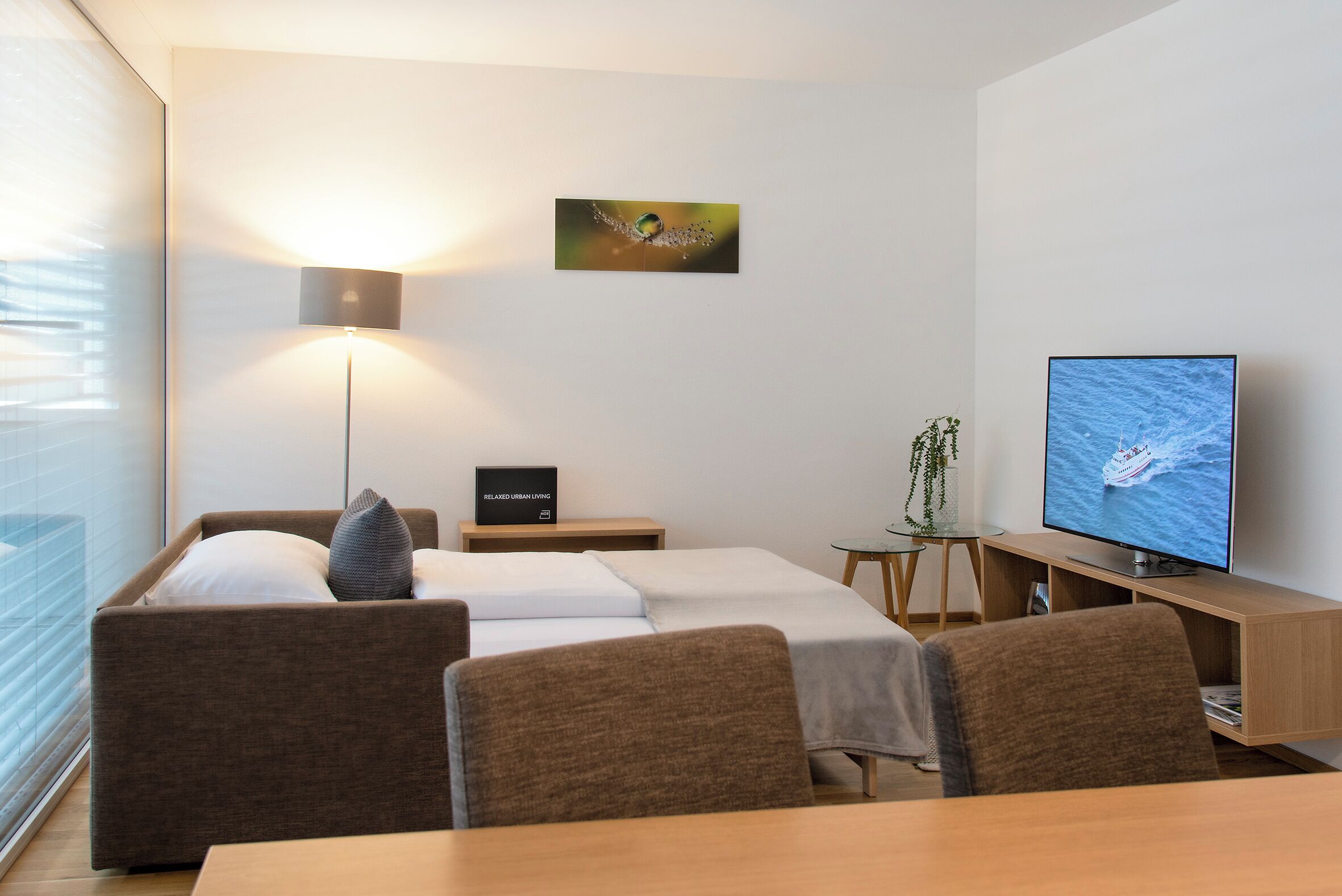 A modern hotel room with a bed, a desk, a television, a lamp, and a small plant.