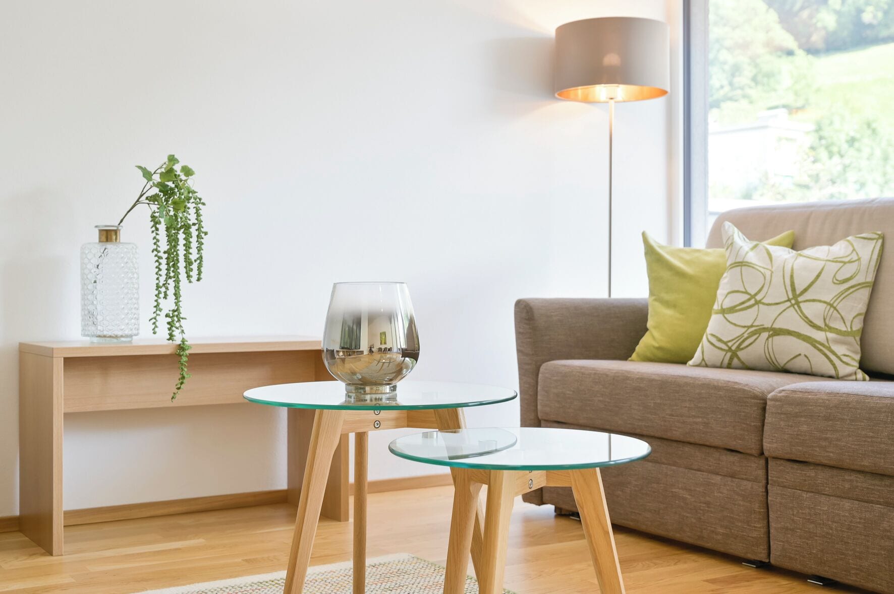 A modern living room with a glass coffee table, a glass wine glass, a hanging plant, a lamp, and a sofa with cushions.
