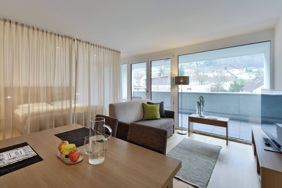 A modern hotel room with a large window, a sofa, a small table, and a dining table with fruits and a glass of water.