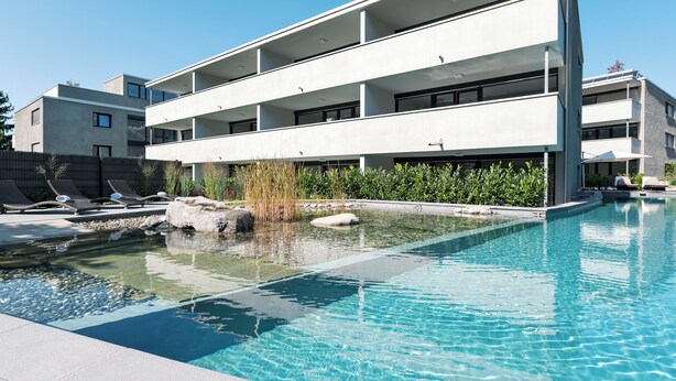 Un edificio moderno con piscina, sillas de descanso y un pequeño estanque.