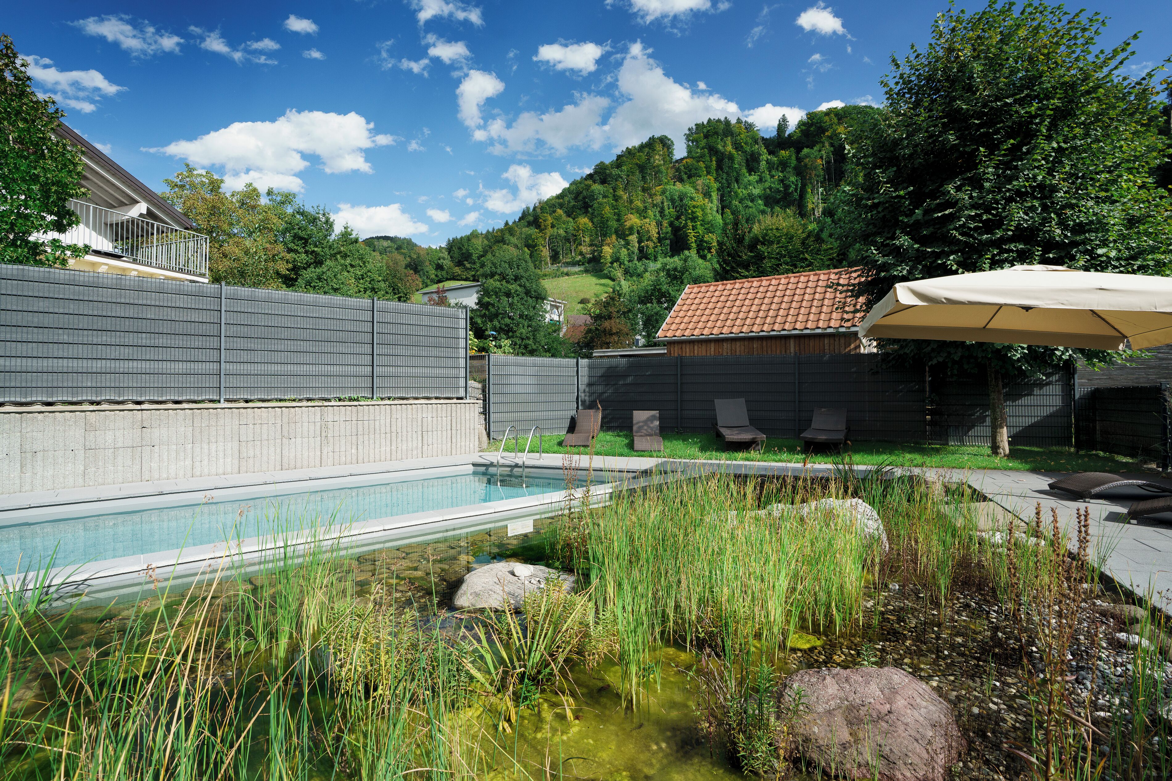 A swimming pool with a tiled deck, surrounded by a fence, and a small pond with rocks and vegetation.
