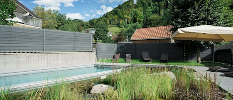 A swimming pool with a tiled deck, surrounded by a fence, and a small pond with rocks and vegetation.