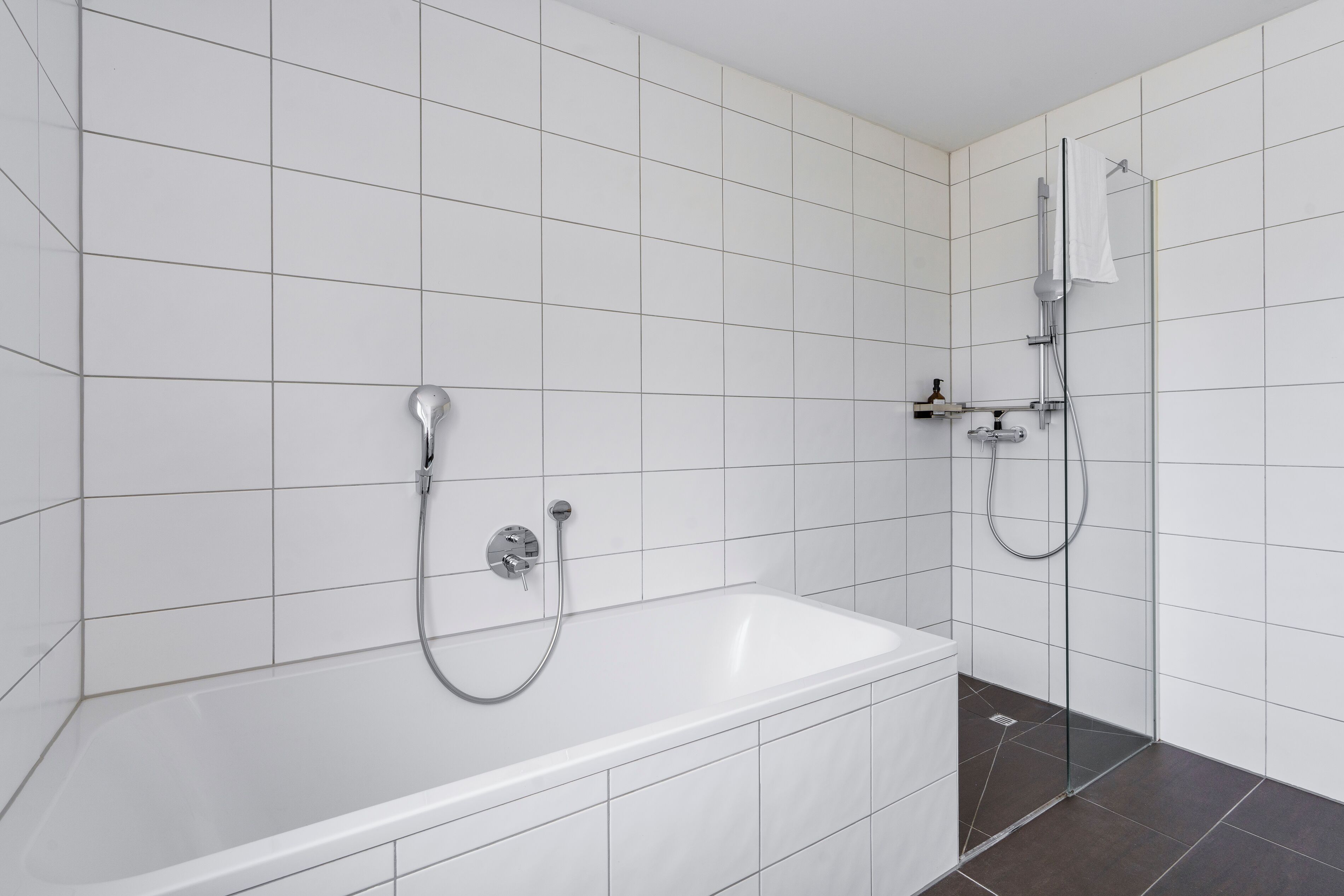 A modern bathroom with a bathtub and shower, white tiled walls, and dark floor tiles.