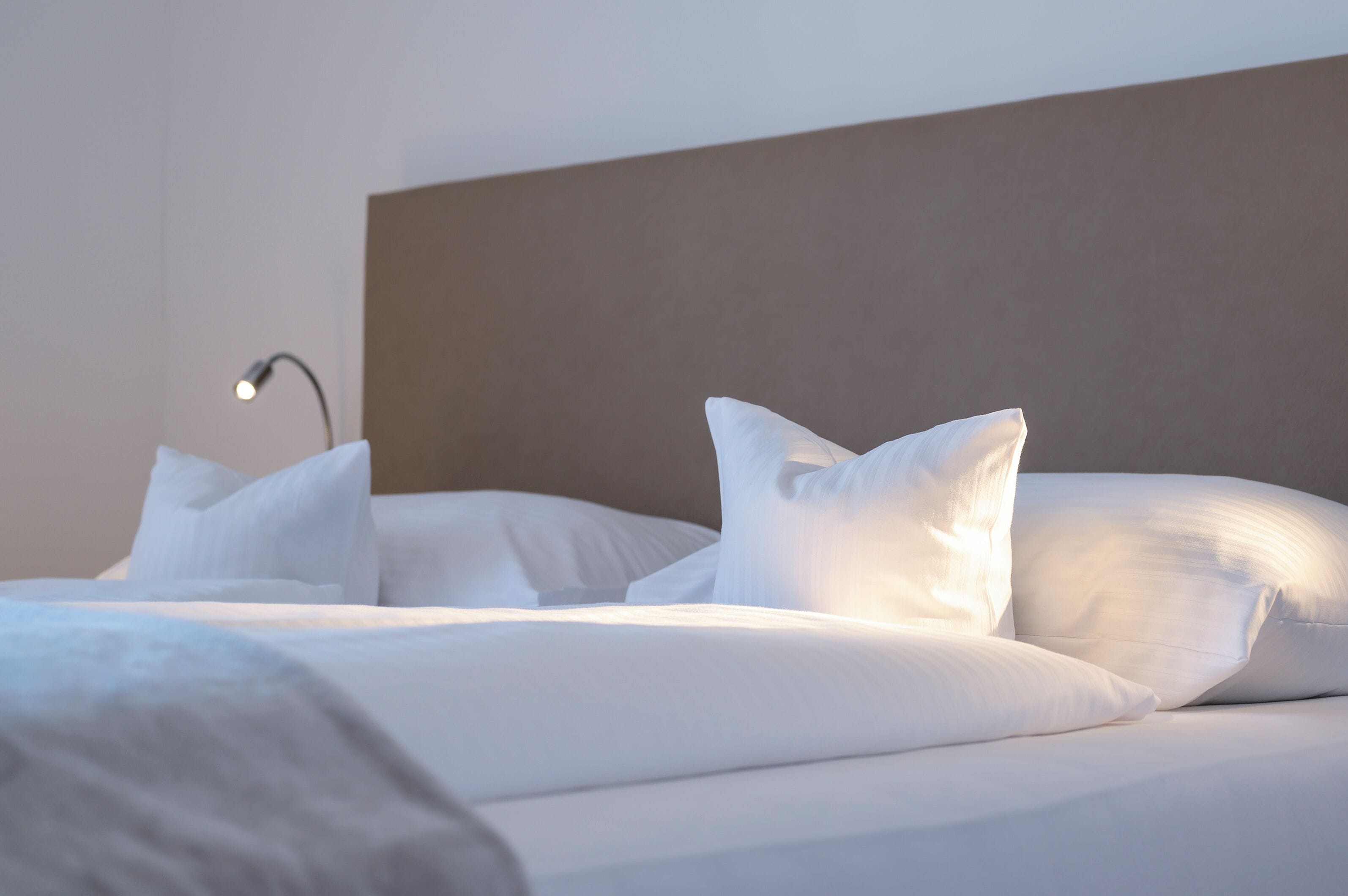 Ein ordentlich bezogenes Bett mit weißen Bettwäsche und Kissen, eine Nachttischlampe und ein schlichter Kopfteil.