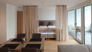 Una habitación de hotel moderna con una cama grande, un área de comedor con mesa y sillas, y un balcón con vistas.