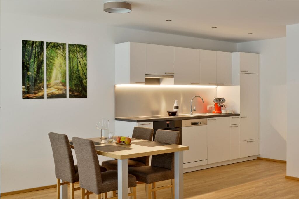 A modern kitchen with white cabinets, a wooden dining table, and a painting of a forest path.
