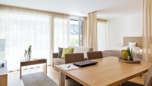 Una habitación de hotel moderna con una mesa de comedor de madera, un sofá con cojines, una mesita auxiliar con una planta y un gran ventanal con cortinas translúcidas.