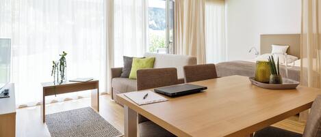 A modern hotel room with a wooden dining table, a sofa with cushions, a small side table with a plant, and a large window with sheer curtains.