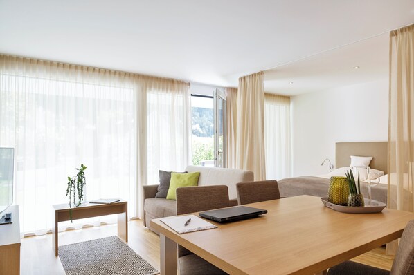 A modern hotel room with a wooden dining table, a sofa with cushions, a small side table with a plant, and a large window with sheer curtains. - Relaxed Urban Living (Dornbirn)