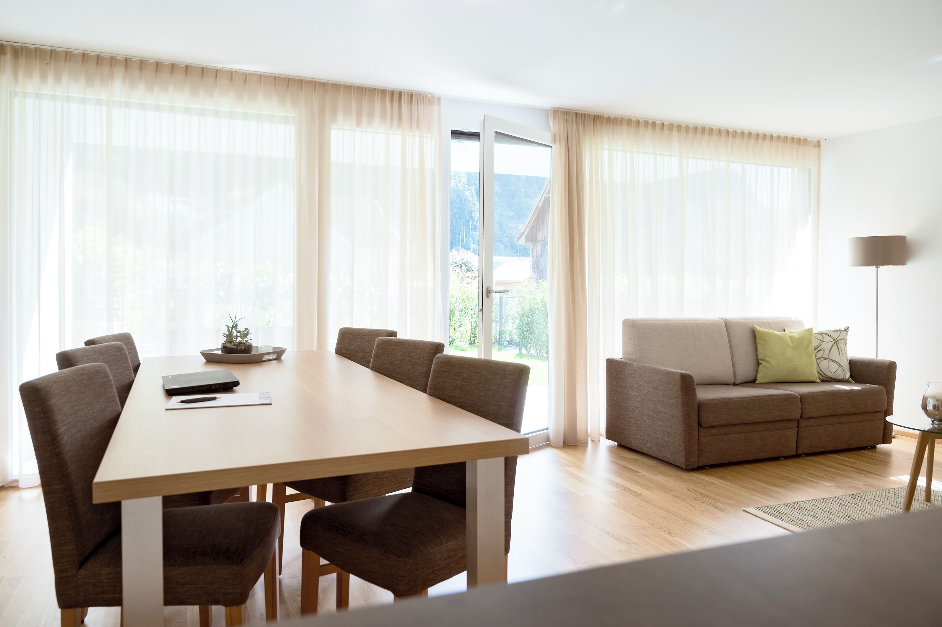 A modern living room with a dining table and sofa, large windows with sheer curtains, and a view of greenery outside.