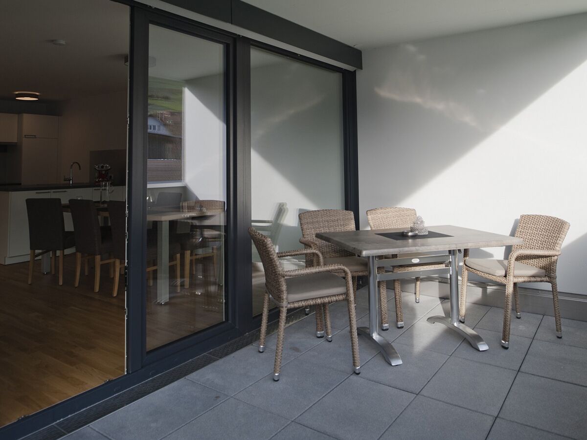 Un área de comedor exterior moderna con sillas de mimbre y una mesa de metal, contigua a un piso corredizo que da a una cocina.