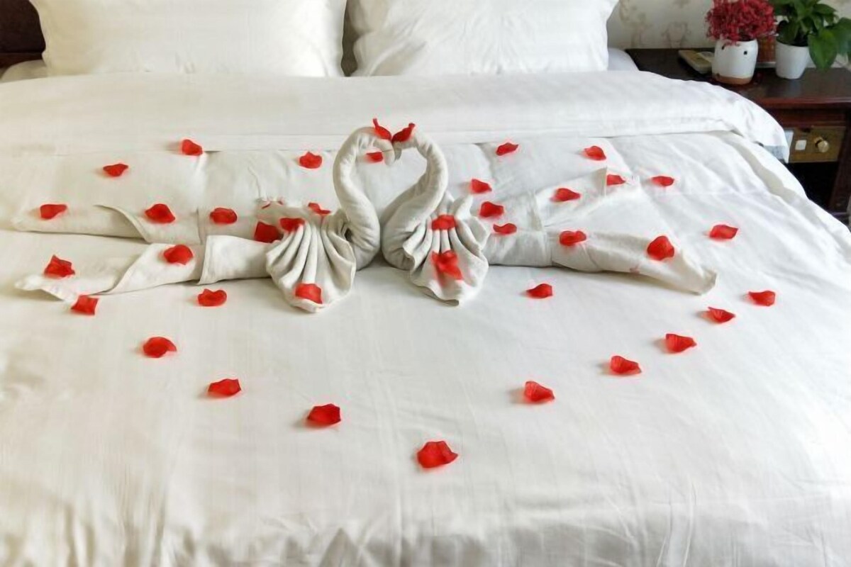 Ein Bett mit weißer Bettdecke, zwei schwanenförmigen Kissen und verstreuten roten Rosenblättern.