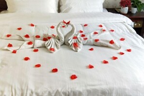Ein Bett mit weißer Bettdecke, zwei schwanenförmigen Kissen und verstreuten roten Rosenblättern.