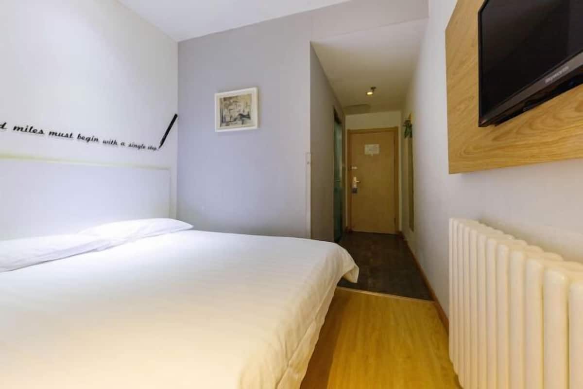 A hotel room with a bed, a wall-mounted TV, a radiator, and a hallway leading to another room.