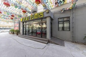 A hotel entrance with a glass facade, a visible entrance door, and a small outdoor seating area with tables and chairs.