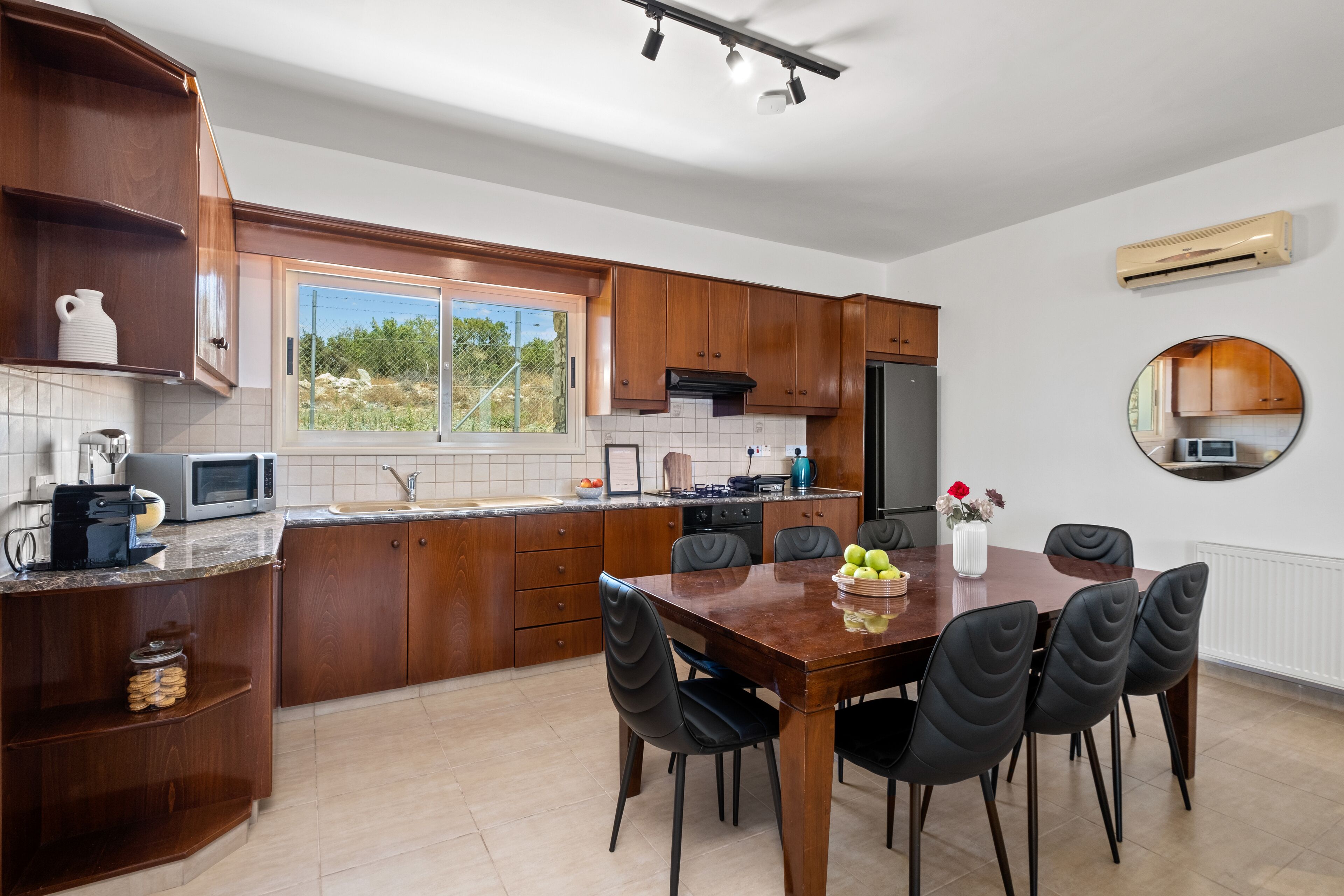 A modern kitchen with wooden cabinets, a dining table with chairs, a microwave, and a window with a view of greenery.