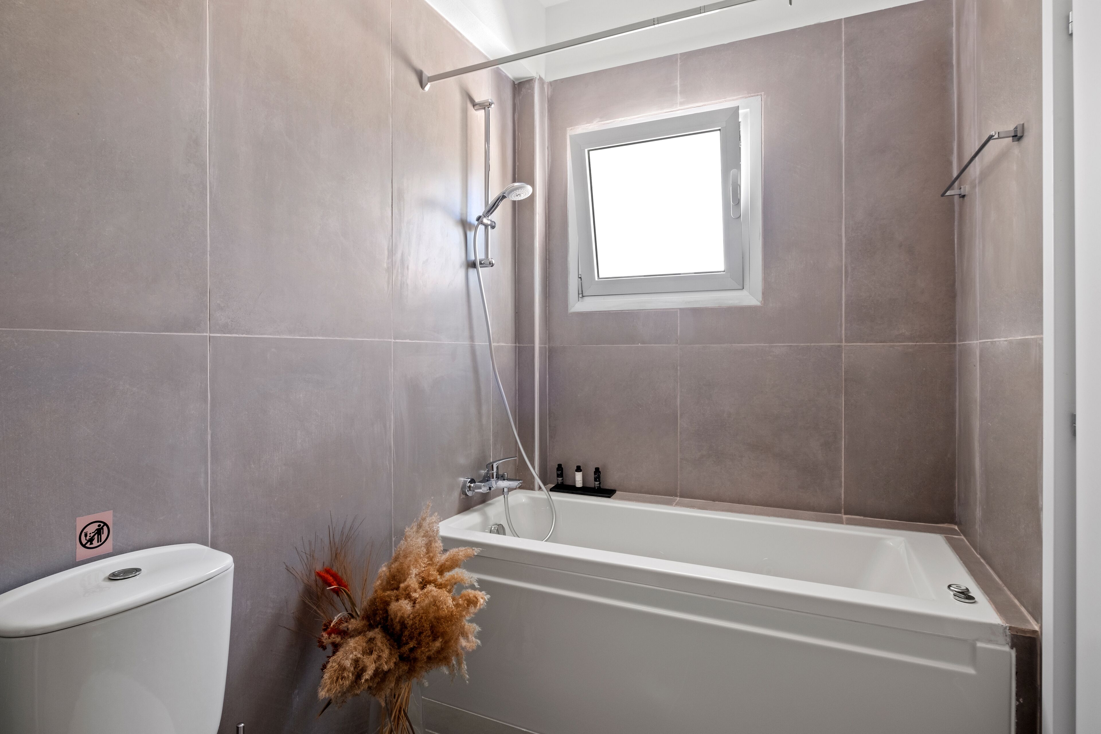 A modern bathroom with a bathtub, shower, and toilet. There is a window providing natural light and a small shelf with toiletries.