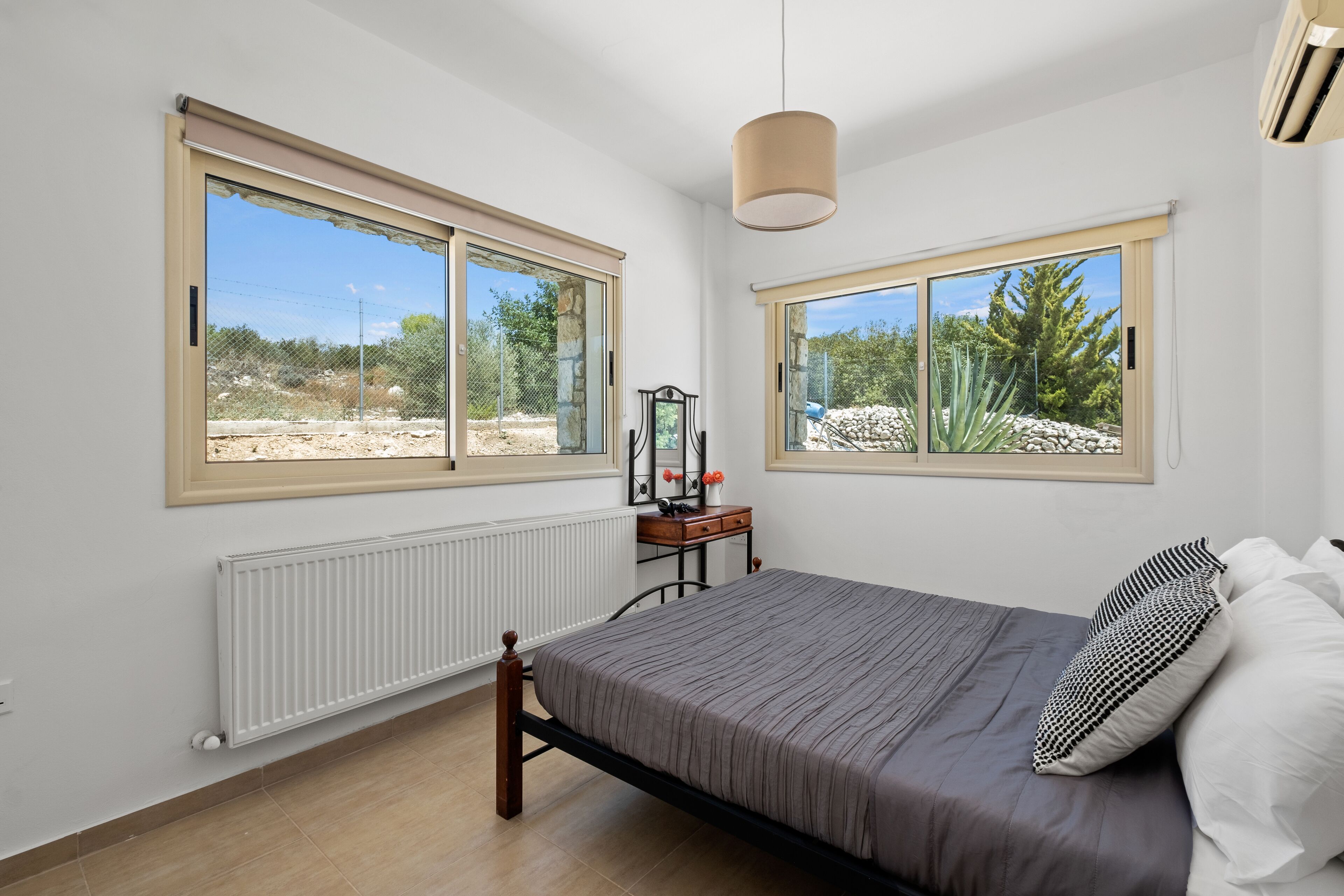 Quarto com cama, duas janelas, radiador e uma pequena mesa com espelho e flores.