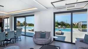 Een moderne woonkamer met een groot raam dat uitkijkt op een zwembad, een ronde grijze bank en een eethoek met stoelen.