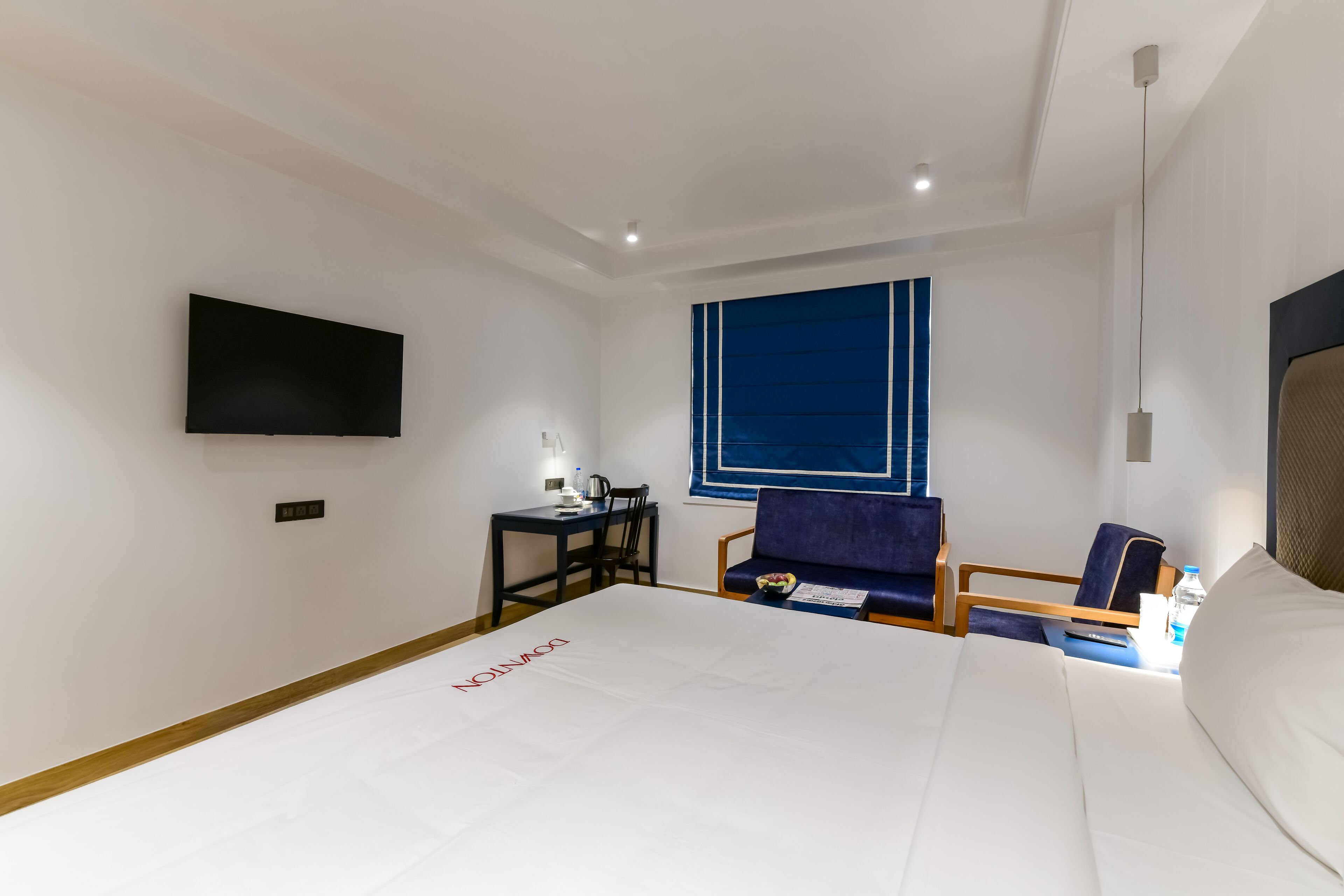 A hotel room with a large bed, a desk with a chair, a TV mounted on the wall, and a window with blue blinds.