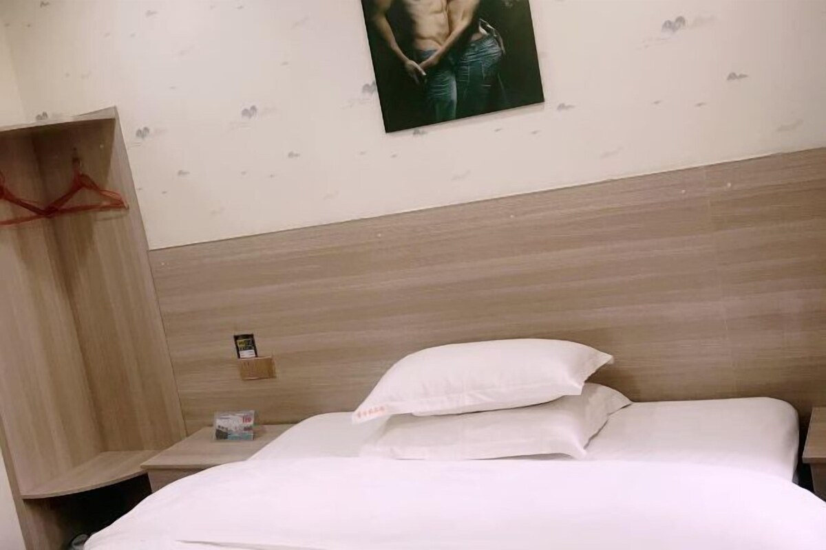 A hotel room with a wooden headboard, a bed with white linens, a small bedside table with a lamp, and a wall-mounted photo.