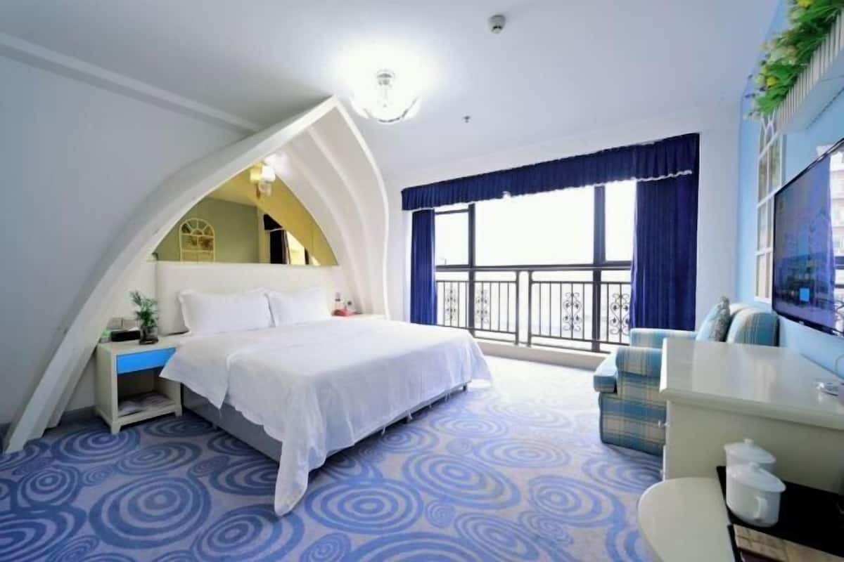 A bedroom with a bed, a small desk, a chair, a TV, and a balcony with a railing.