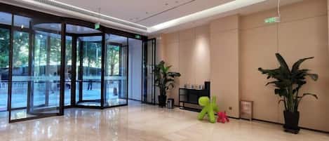 A modern lobby with glass doors leading to an outdoor area, a reception desk, and potted plants.