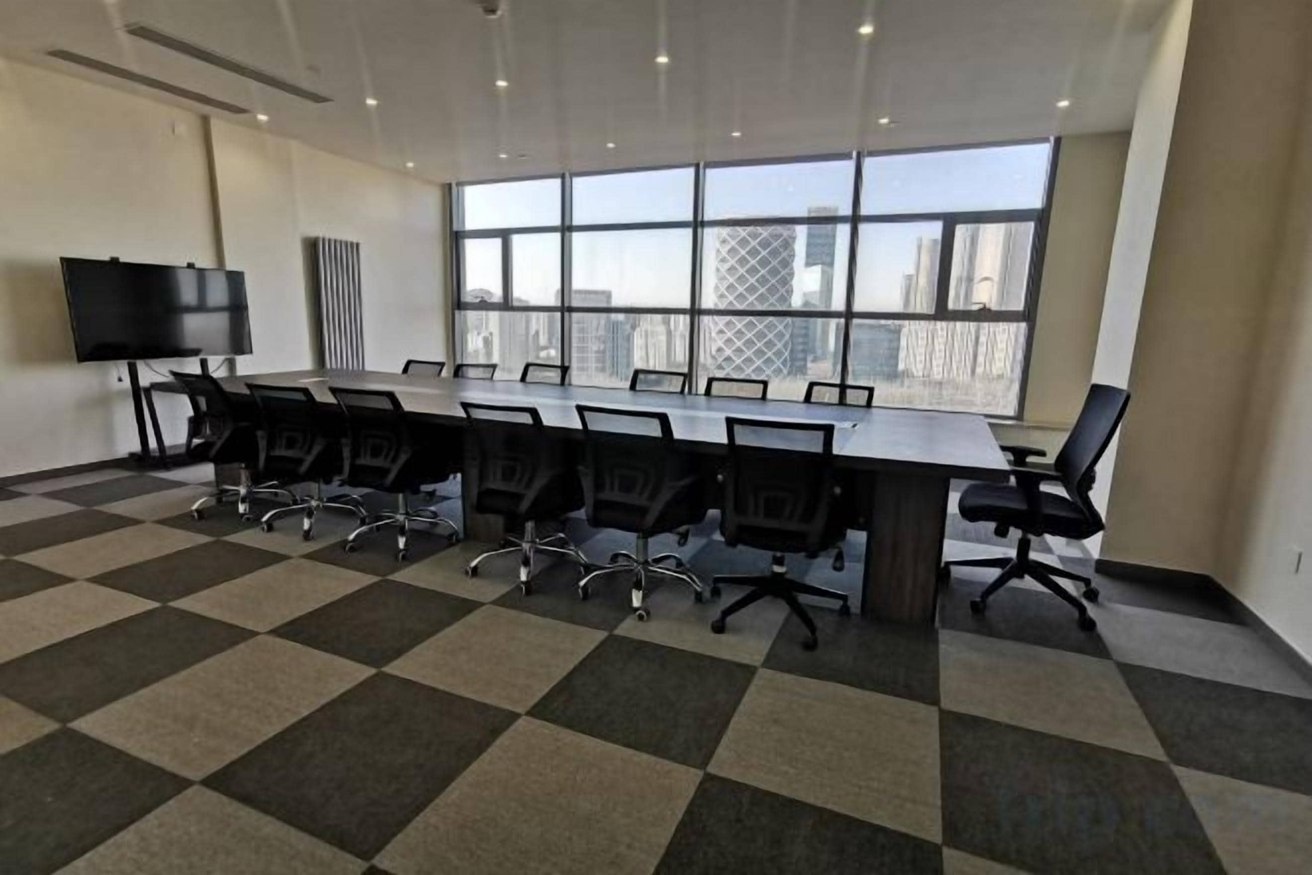 A conference room with a long table, chairs, a TV screen, and a large window with a city view.