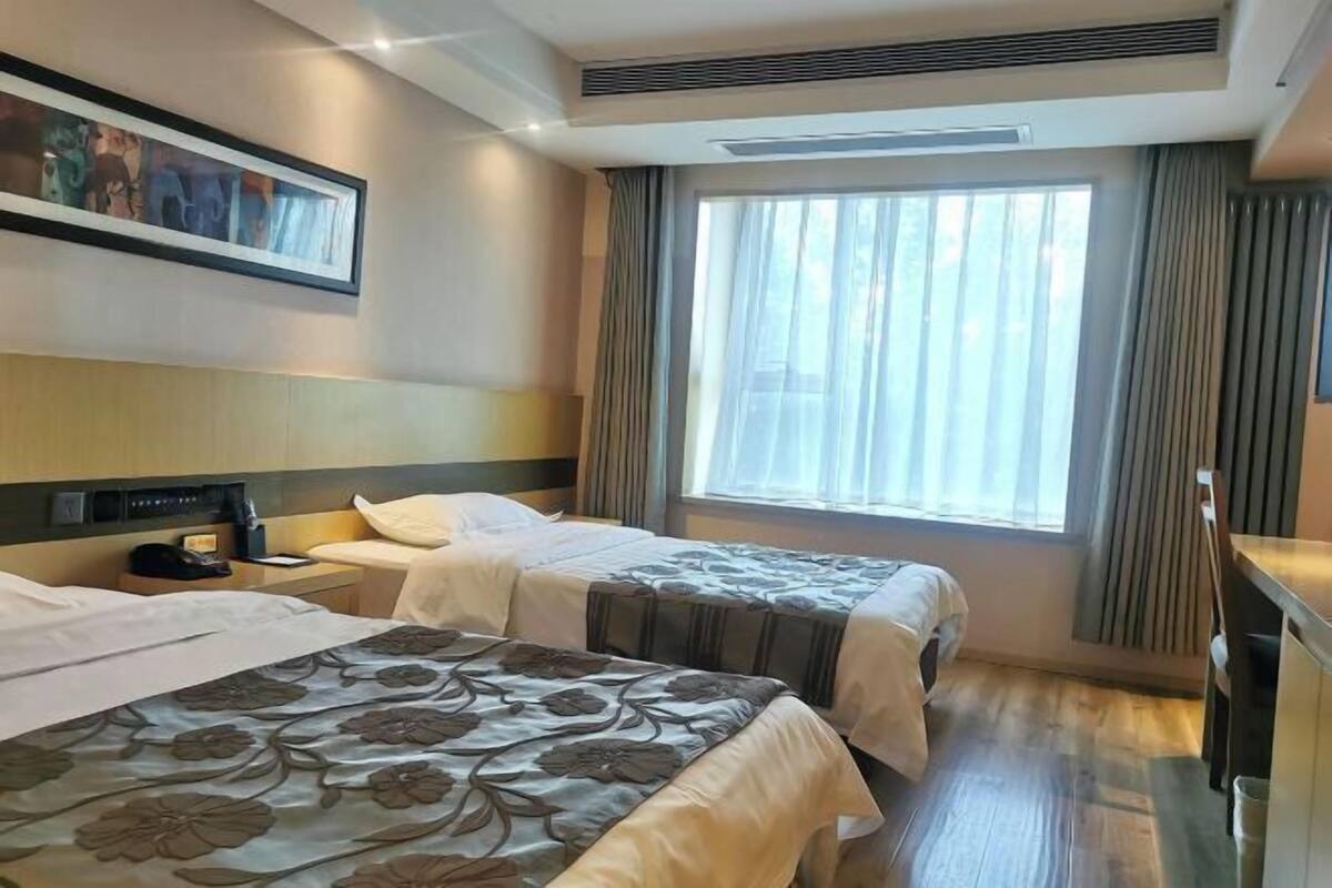 Une chambre d’hôtel avec deux lits, une grande fenêtre avec des rideaux, une tête de lit et un tableau encadré au mur.