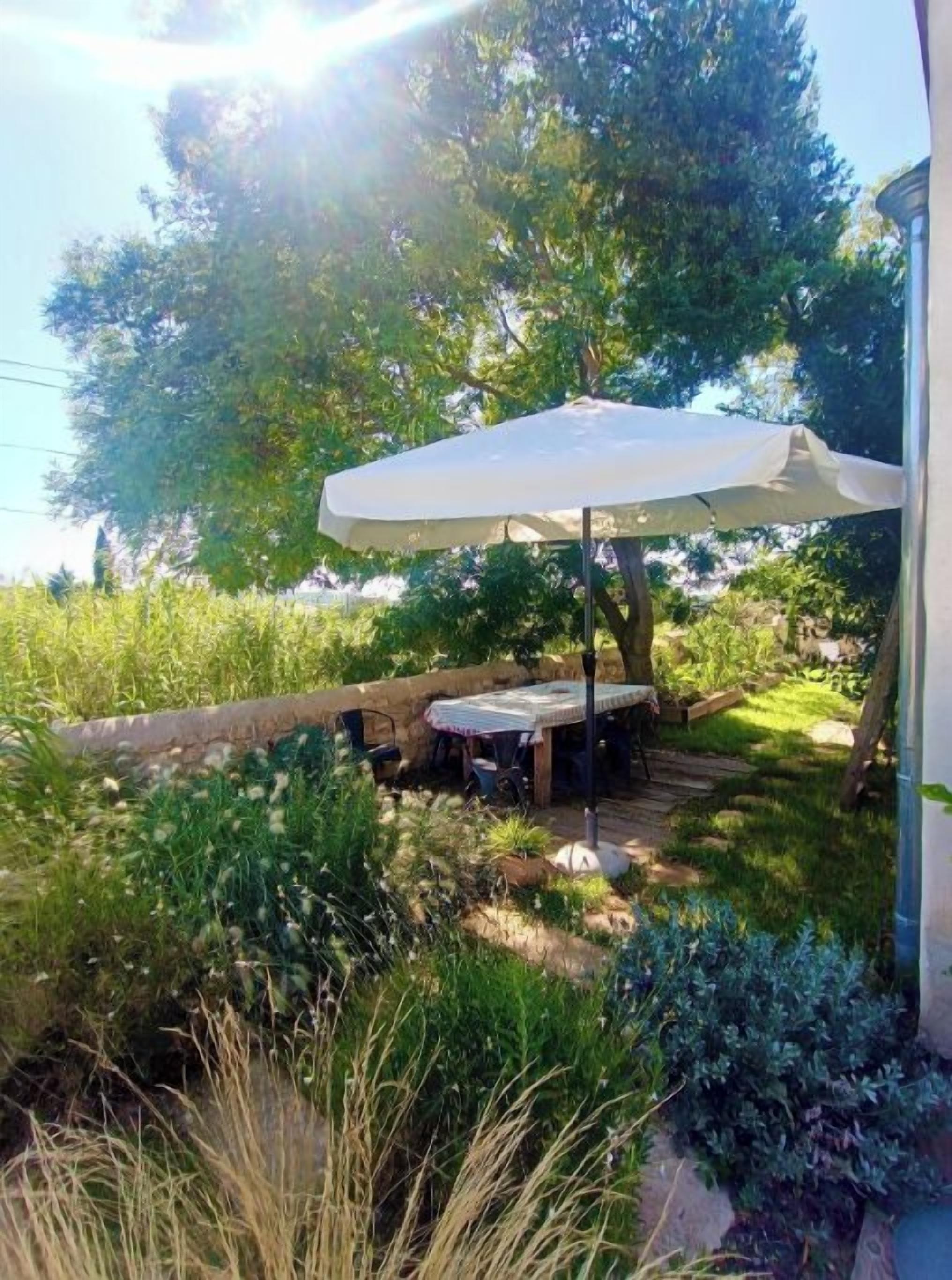 Un patio al aire libre con una mesa y sillas bajo un paraguas blanco, rodeado de follaje y un muro de piedra.