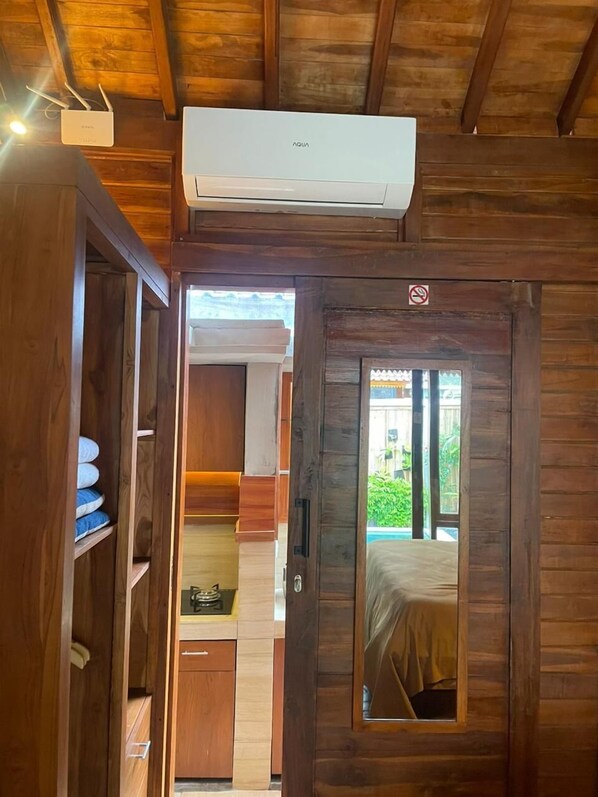 A wooden interior with a central air conditioning unit, a door with a no-smoking sign, and a glimpse of a bed through a window. - Ngesil Private Joglo and Pool (Ubud)