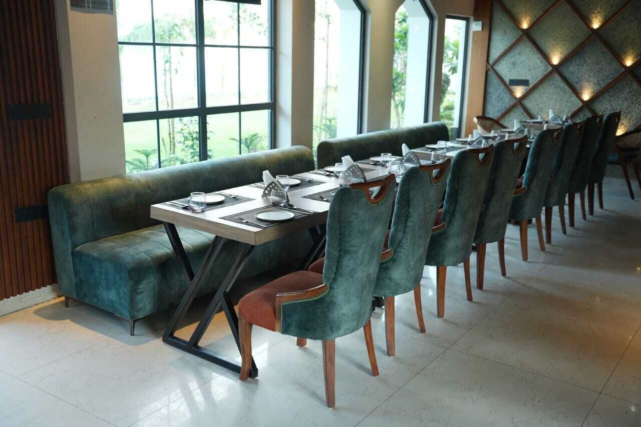 A dining area with a long table set for multiple people, cushioned seating, and large windows offering a view of greenery outside.